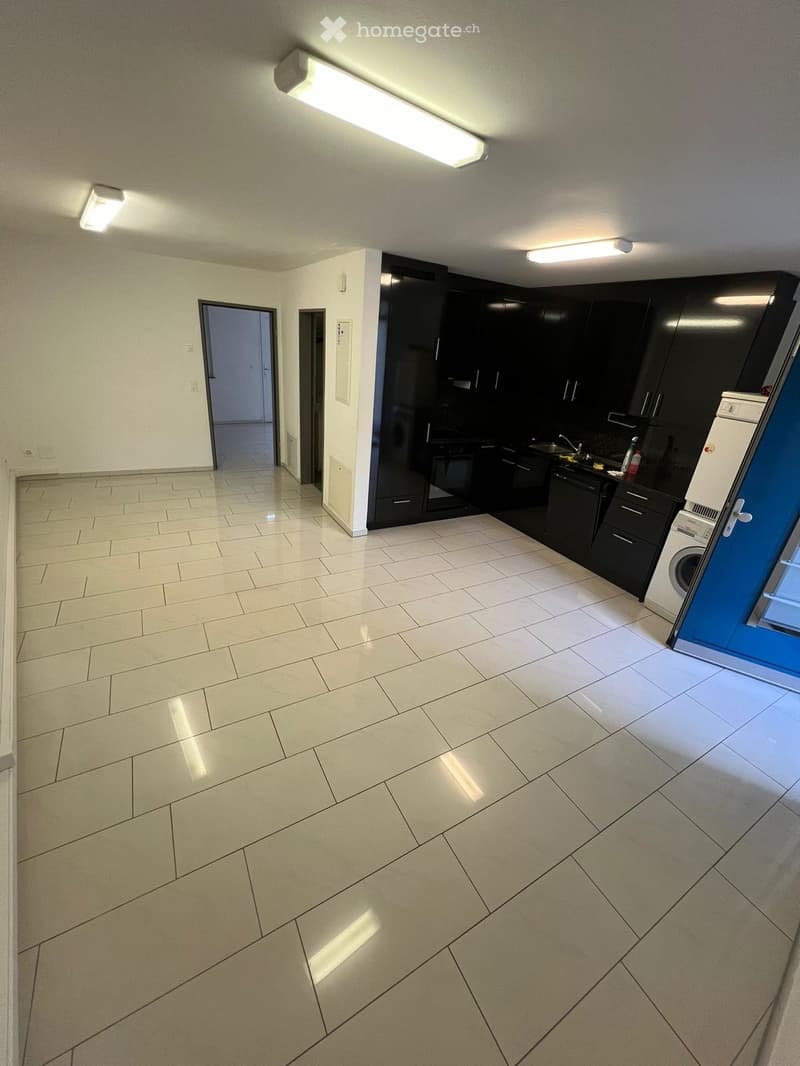 Empty room with tiled floor, white walls, and a small kitchen area with black cabinets and a washing machine visible in the background.