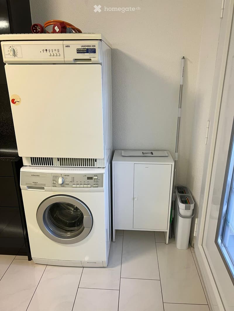 A small bathroom with a white washing machine, a white dishwasher, and a white trash bin. The floor is tiled, and the walls are light-colored.