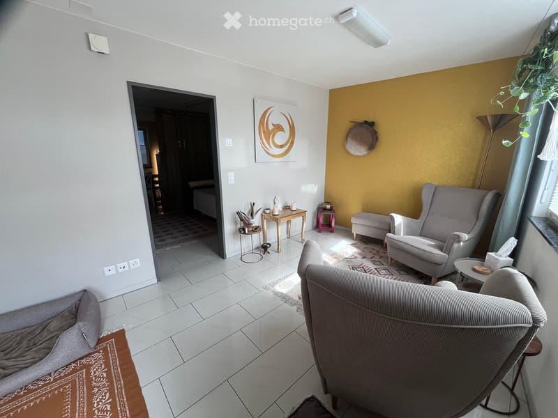 Modern living room with grey sofa, armchair, small tables, floor lamp, and decorative items. The space features a mix of light and dark walls, a patterned rug, and a view into an adjacent room.