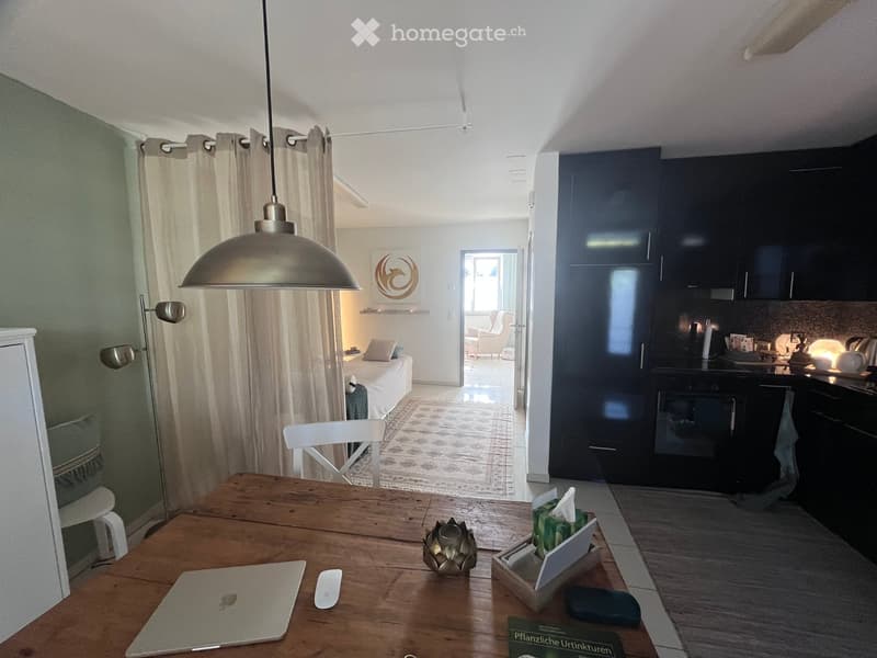 A modern, open-concept living space with a wooden dining table, laptop, pendant light, and a visible bedroom area with a bed and pillows. The kitchen features dark cabinets and an oven.