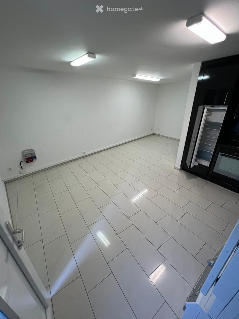 Empty room with white tiled floor and walls, fluorescent ceiling lights, and a partially visible refrigerator in the background.