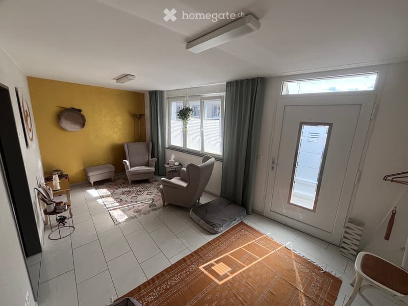 Bright living room with tiled floor, large window, grey armchairs, small table, and decorative items.