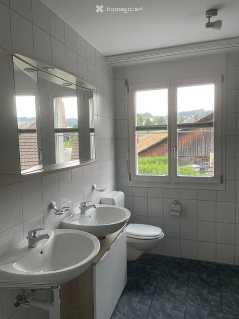 Modern bathroom with two sinks, a toilet, a large mirror, and a window offering a view of the outdoors.