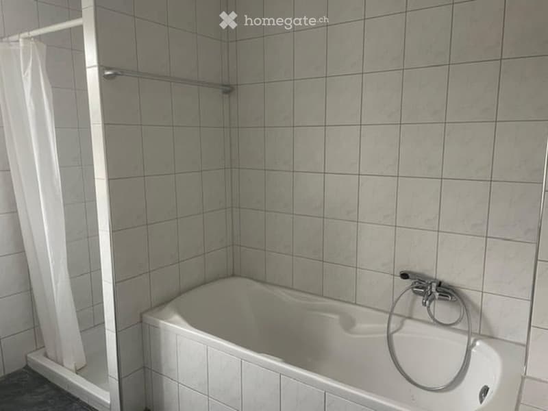 A simple bathroom with a white bathtub, a shower curtain, and a shower rail. The walls are tiled with light grey tiles.