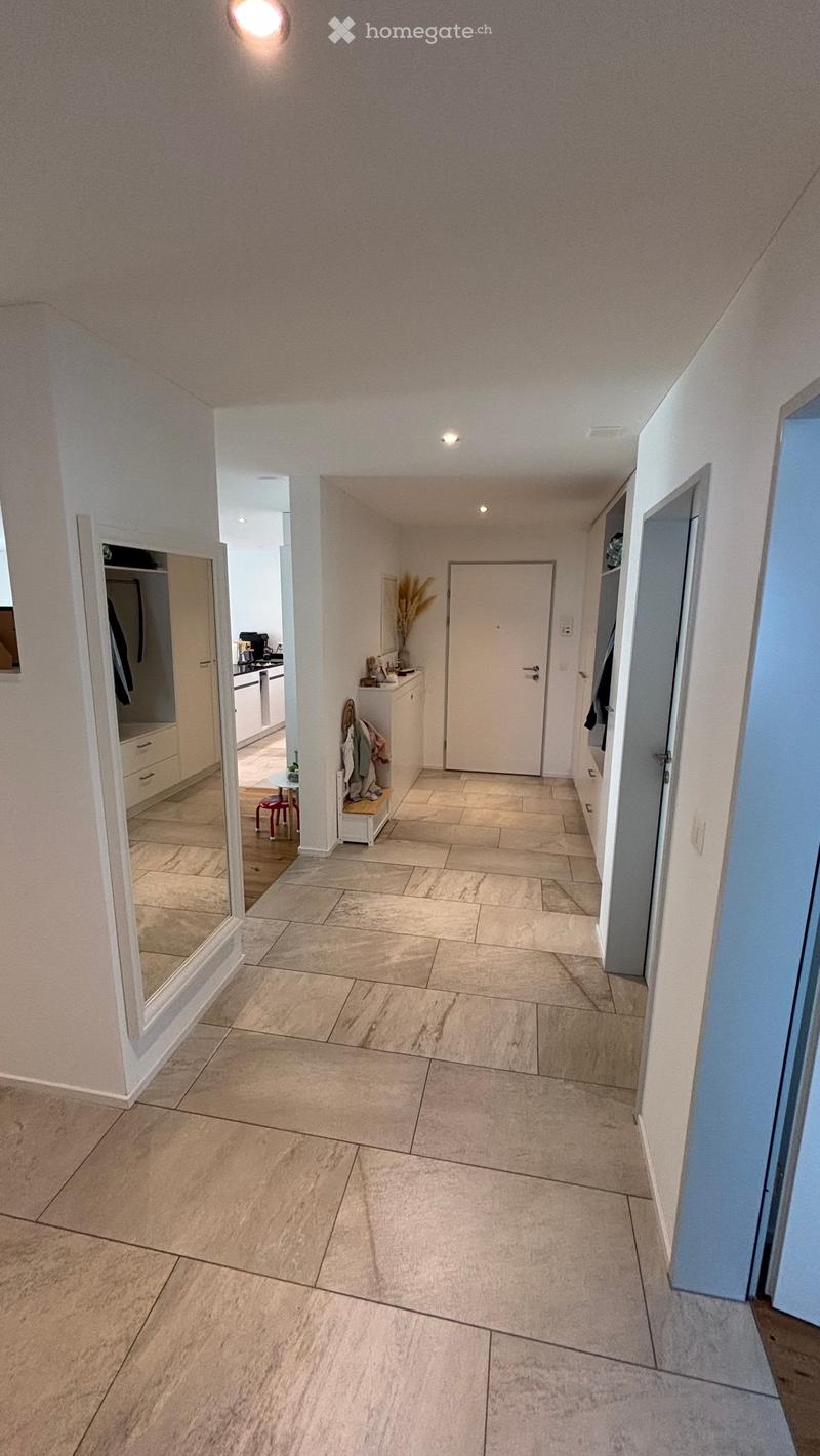 Clean, modern hallway with tiled flooring, white walls, and recessed lighting. Features a large mirror on the left, a small table with a chair, and a coat rack with jackets and a vase of dried plants.