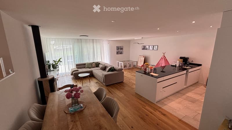 Open-plan living and dining area with a grey sectional sofa, wooden dining table with chairs, kitchen island, and a small playpen. The space features wooden flooring and large windows with sheer curtains.