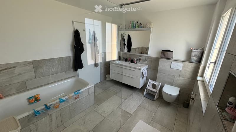 Modern bathroom with a bathtub, sink, toilet, mirror, and a glass shower enclosure. Includes a bathrobe hanging on the wall and a small stool near the toilet.