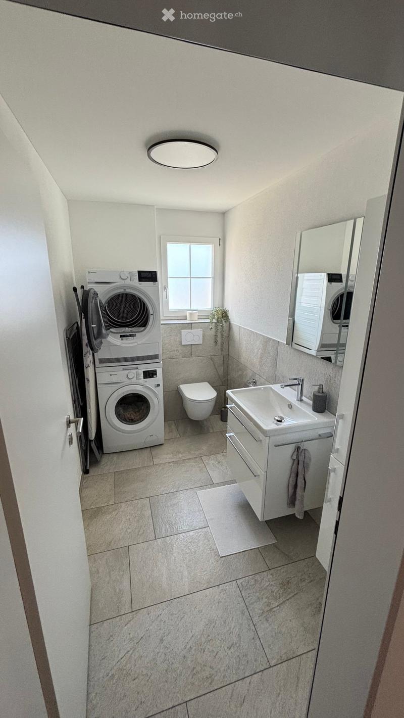 Modern bathroom with a sink, toilet, washing machine, and a mirror. The space features light-colored tiles and a window for natural light.