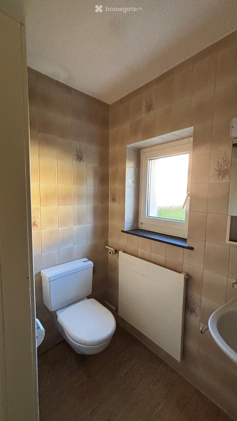 Bathroom with beige tiled walls, a toilet, a sink, a radiator, and a window with a small shelf above it.