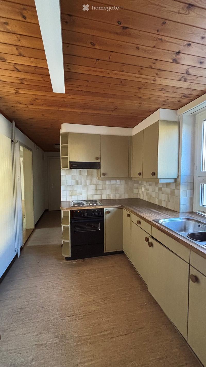 Empty kitchen with light-colored cabinets, a stove, an oven, a sink, and a range hood. The walls are tiled, and the floor is carpeted.