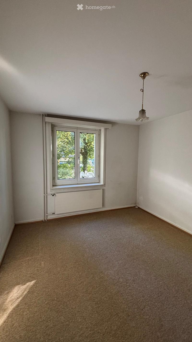 Empty room with beige carpet, white walls, a window with white frame, radiator, and a hanging light fixture.