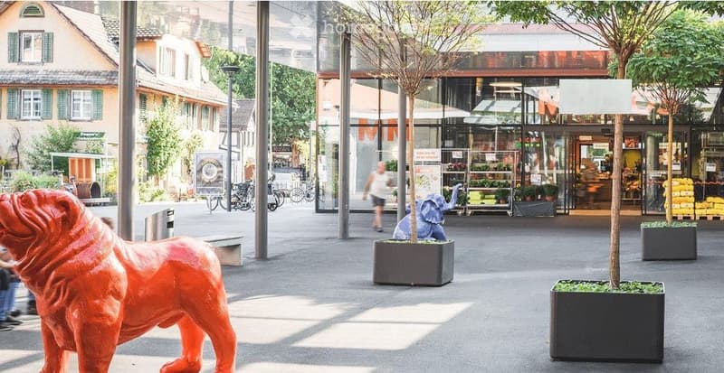 Public square with modern sculptures, trees in planters, and a store in the background.
