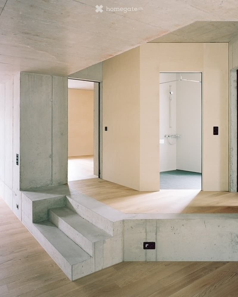 Minimalist, unfurnished room with exposed concrete walls and wooden flooring. Features a small staircase leading to an open area and a visible bathroom with white tiles.