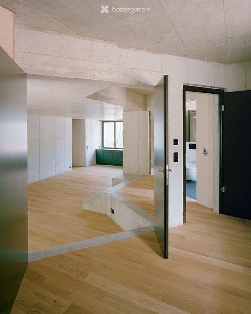 Open, minimalist interior with light wooden flooring, white walls, and visible rooms including a kitchen area and a bathroom. No furniture or personal items are present.