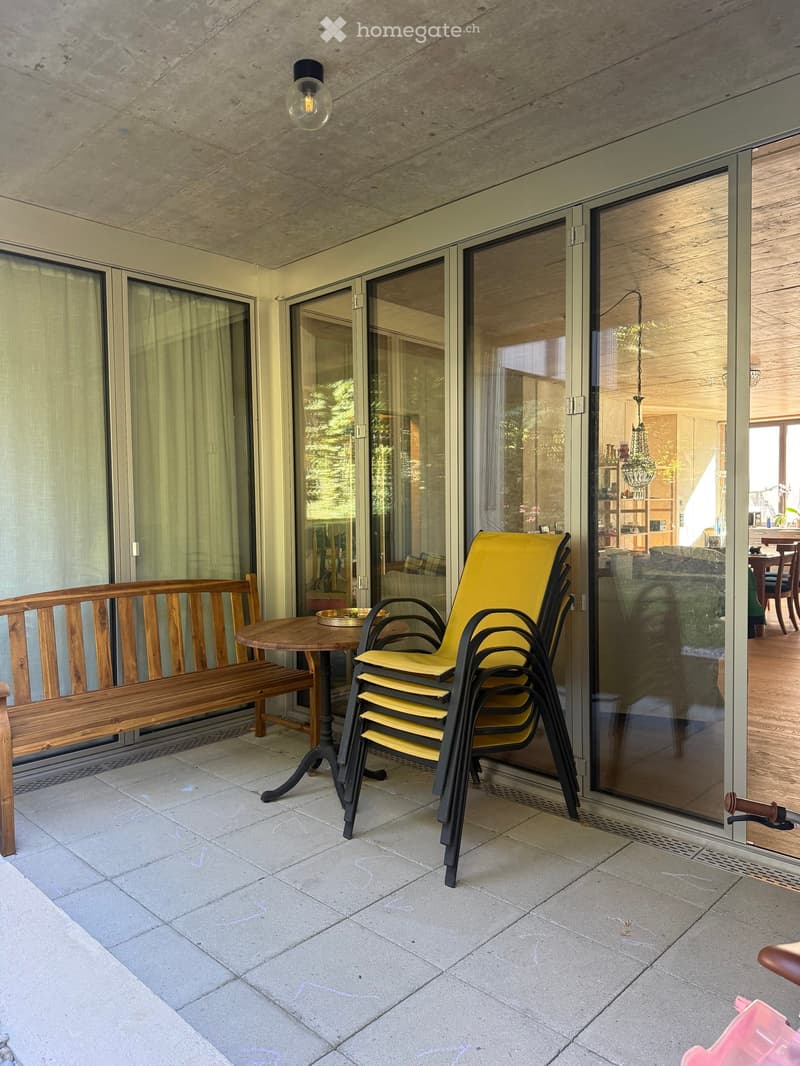 Balcony with a wooden bench, a round table, and a stack of yellow chairs. Large glass sliding doors lead to the interior.