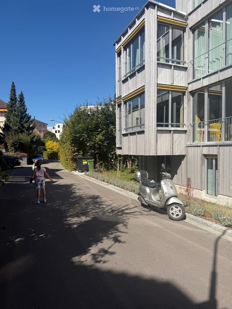 Modern multi-story building with wooden facade, large windows, and balconies. A scooter is parked in front, and a child is walking on the sidewalk.