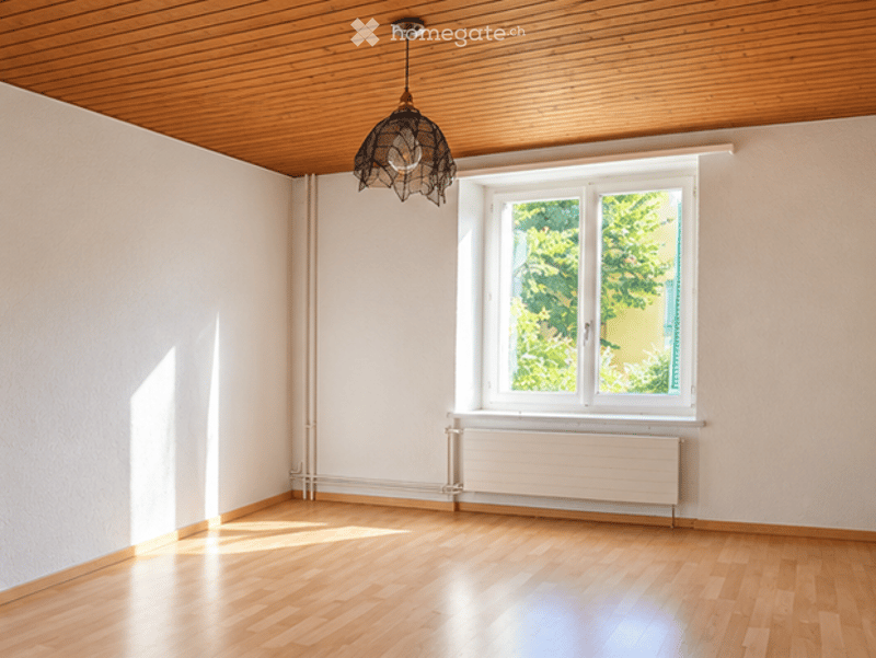 Empty room with wooden flooring, white walls, large window with view of greenery, radiator below window, and a hanging light fixture.