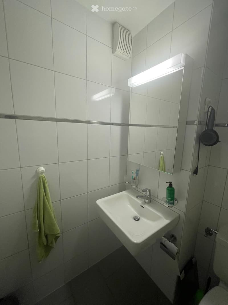 Clean bathroom with white tiled walls, a white sink with a chrome faucet, a mirror with built-in lighting, a towel hanging on the wall, and a soap dispenser on the sink counter.