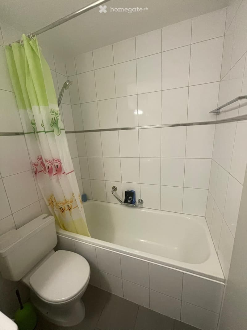 Bathroom with white tiled walls and floor, featuring a toilet, bathtub, and a shower curtain with cartoon characters.