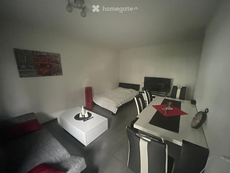 A compact, furnished room with a bed, sofa, coffee table, dining table, chairs, and a TV screen. The room features a modern design with a mix of white and dark furniture.