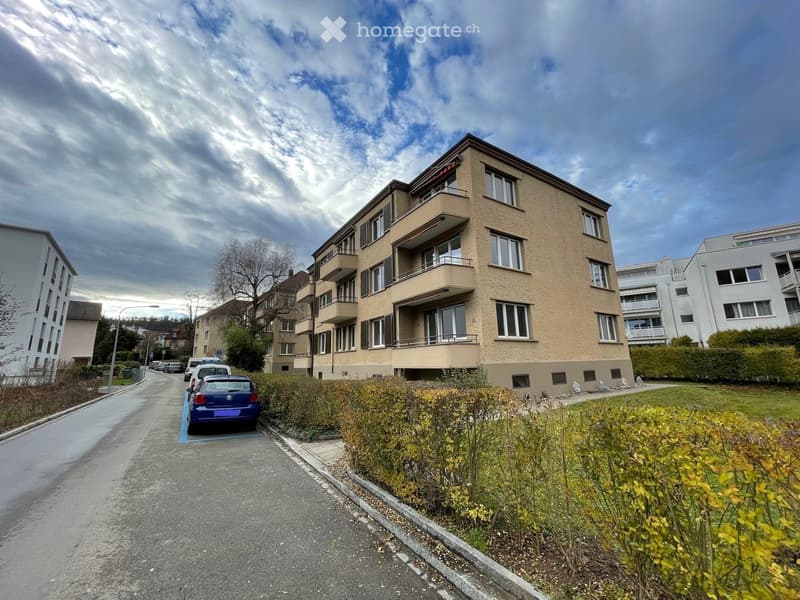 Multi-story residential building with balconies, surrounded by greenery and parked cars.