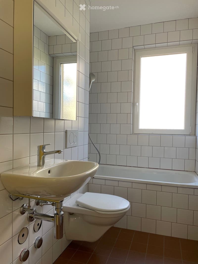 Modern bathroom with white tiled walls, a sink with a chrome faucet, a toilet, a bathtub, and a large mirror above the sink. Natural light enters through a window.