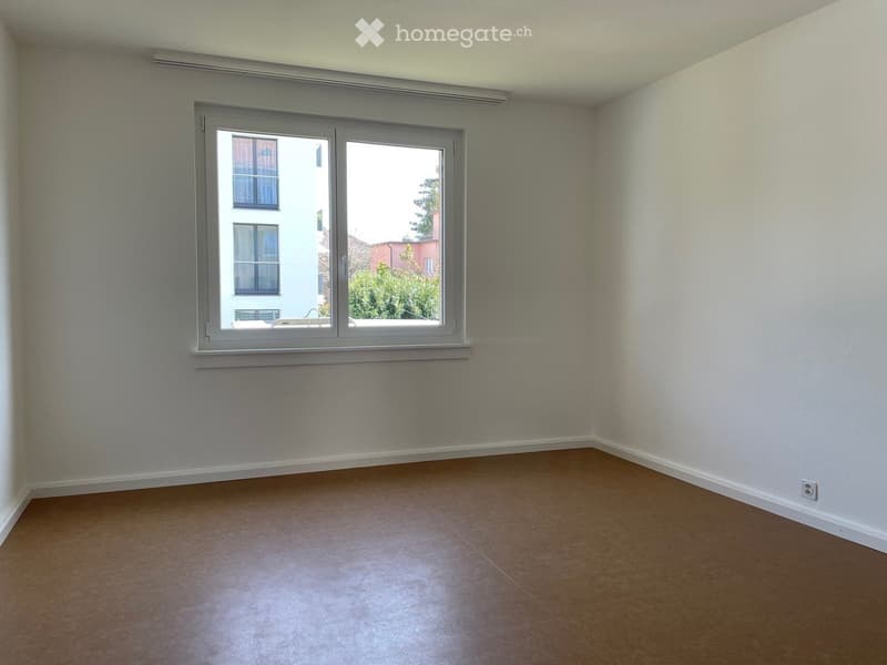 Empty room with light-colored walls, tiled floor, and a large window offering a view of neighboring buildings and greenery.
