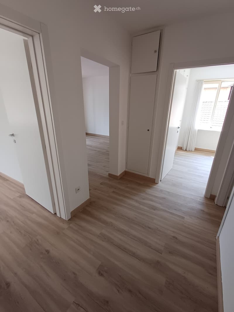 Empty hallway with light wooden flooring and white walls, featuring several doors and a window with curtains at the end.