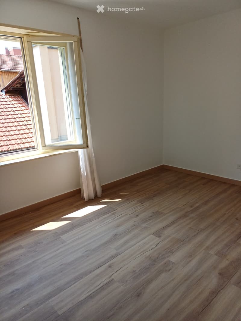 Empty room with wooden flooring and large window showing a neighboring roof. Bare walls with no furniture or decorations.