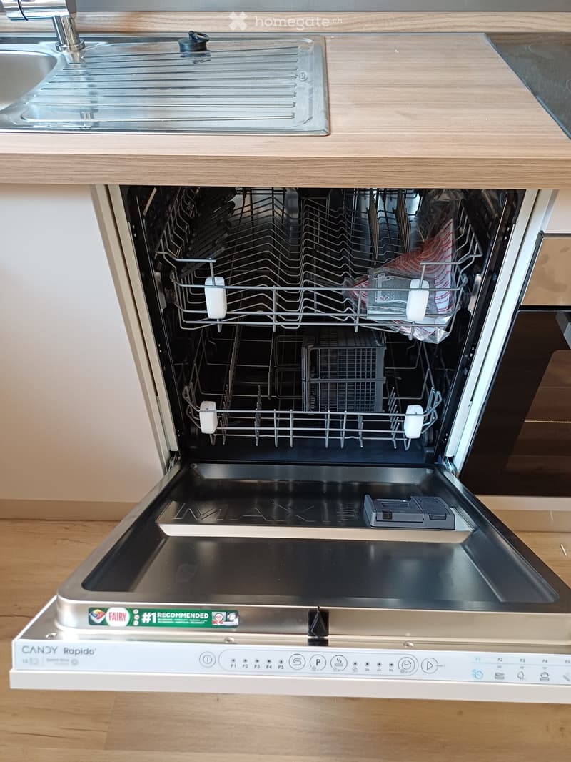 Open stainless steel dishwasher with multiple racks and control panel, placed under a wooden countertop with a sink visible above.