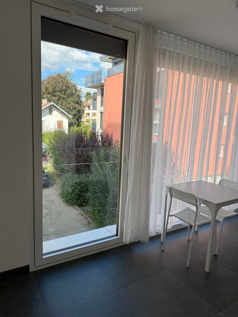 A modern balcony with a sliding glass door, white curtains, and a small dining setup with a table and chairs.