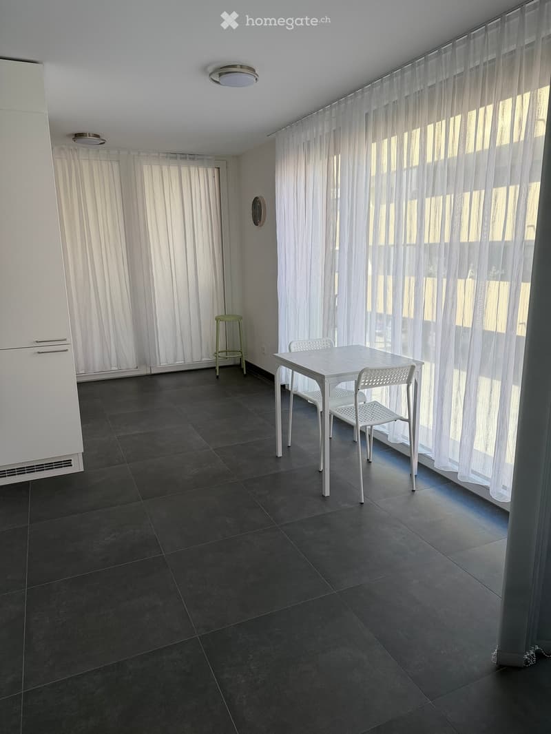 Bright room with dark tiled floor, white table and chairs, green stool, large windows with white curtains, and a white cabinet.