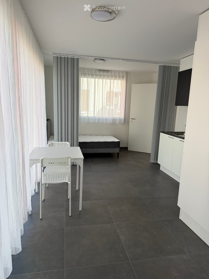 A modern, open-concept room with a bed, a small dining table with chairs, and a compact kitchen area with white cabinets and a sink.