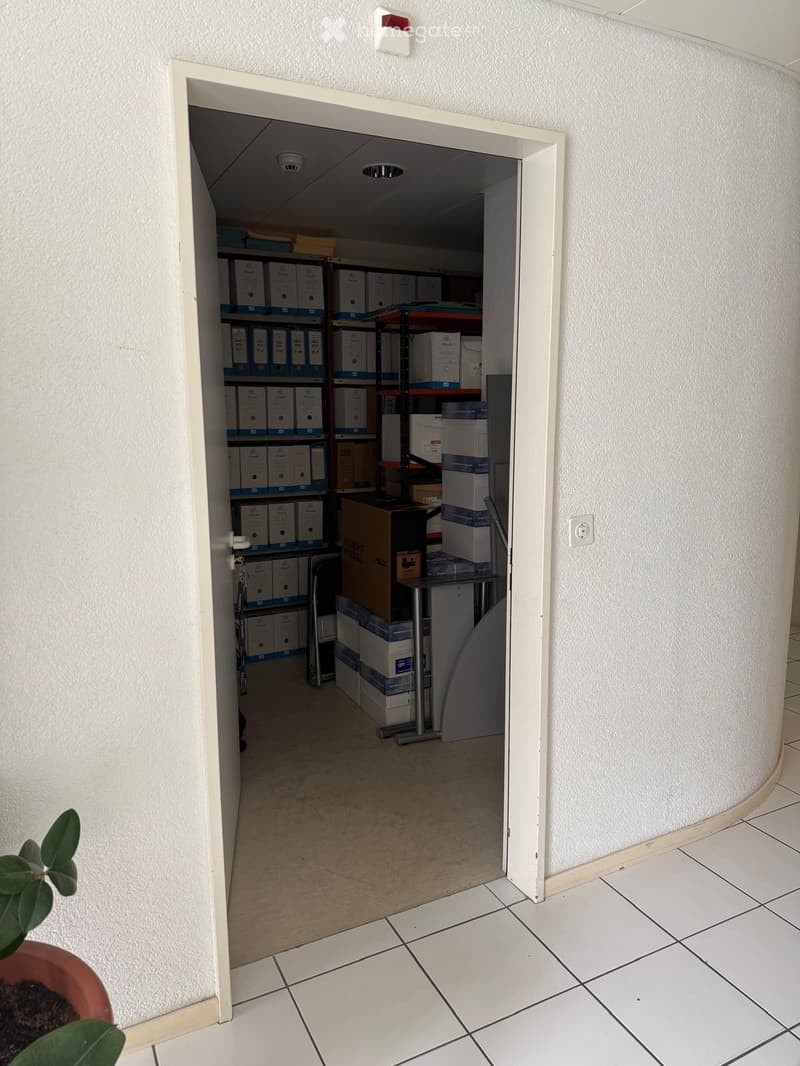 A narrow hallway with white tiled flooring and walls, leading to a storage area filled with boxes and shelves. The storage area contains various cardboard boxes and storage bins arranged on metal shelving units.