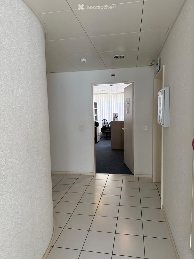 Clean, tiled hallway with white walls, a door leading to an office area, and a security panel on the right wall.