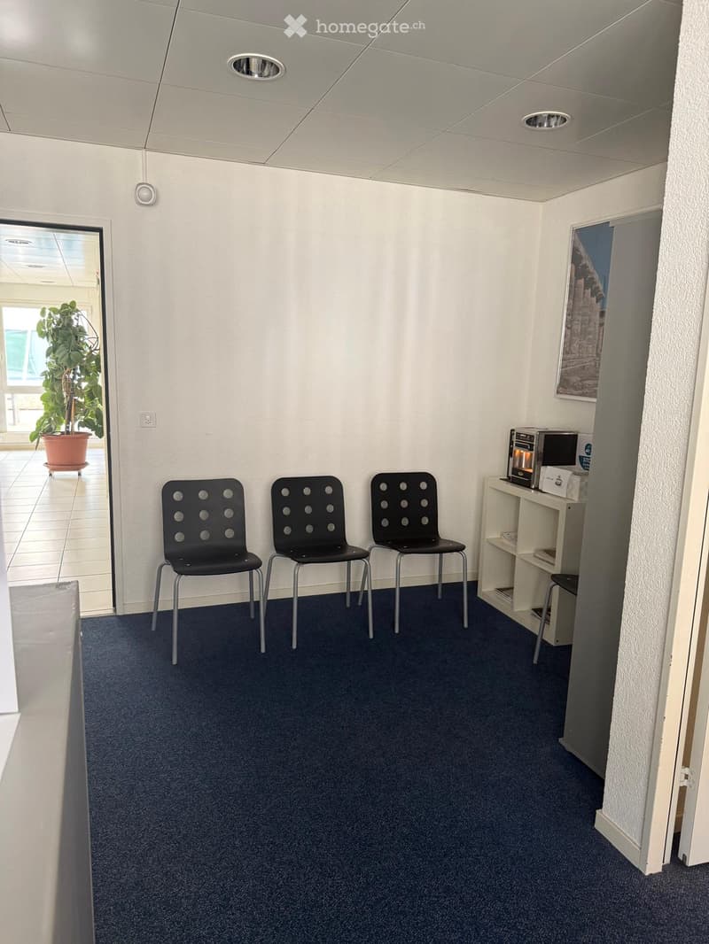 A small, furnished room with three black chairs, a small table with a microwave, a stool, and a picture frame on the wall. The room has blue carpeting and white walls.
