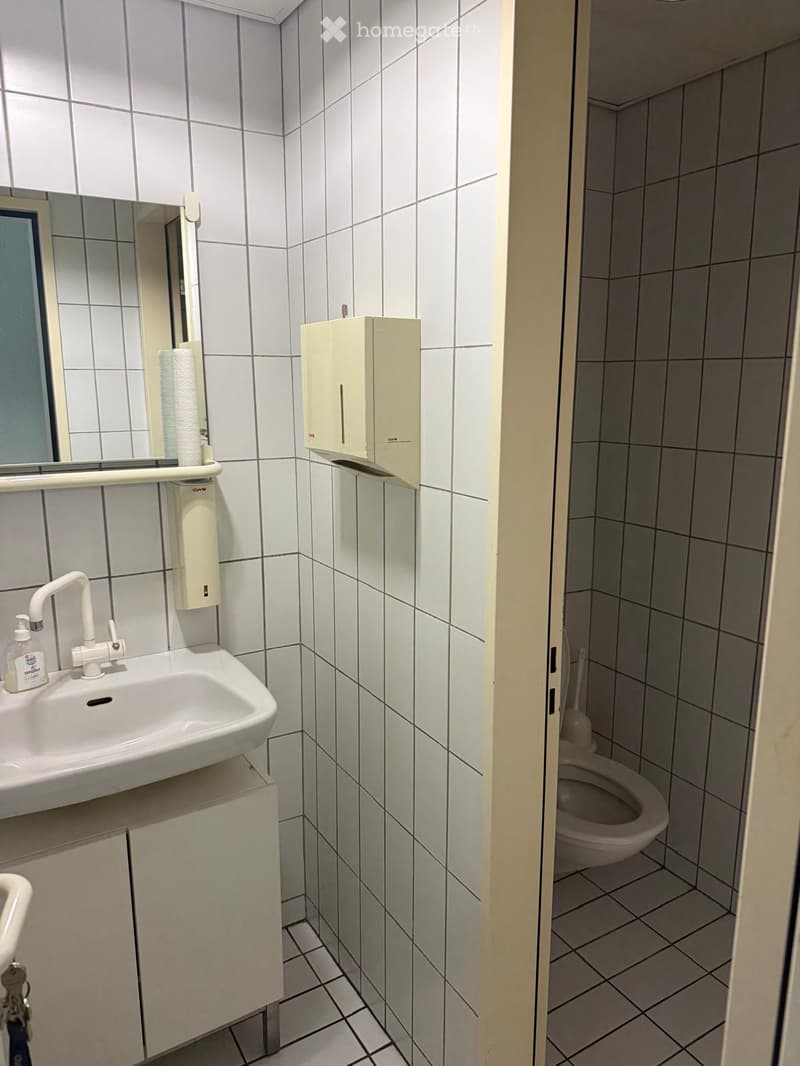 Small bathroom with white tiled walls and floor, featuring a sink with a mirror, a toilet, and soap and paper towel dispensers.