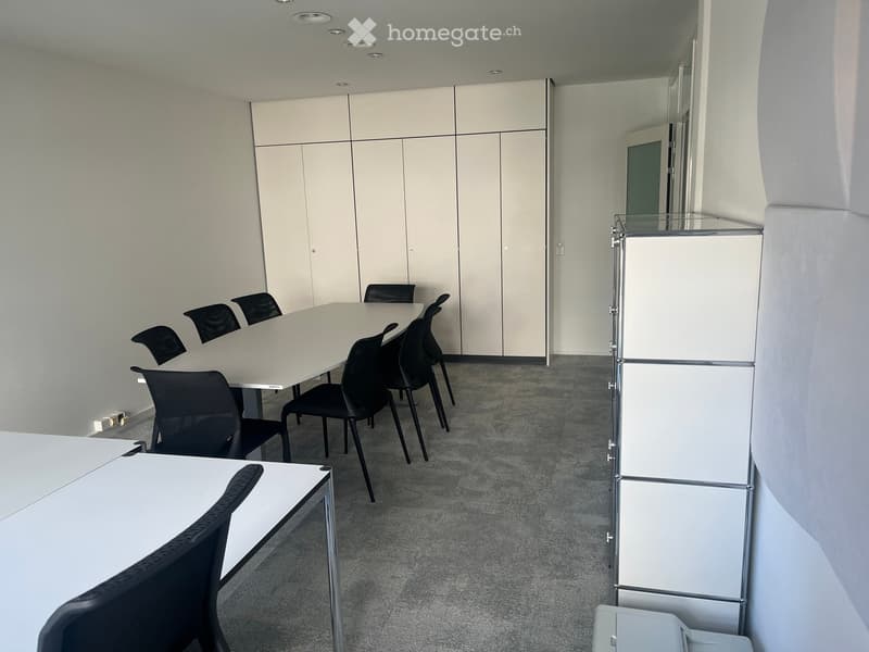 Modern office space with white walls, grey carpet, white tables, and black chairs arranged around a central table.