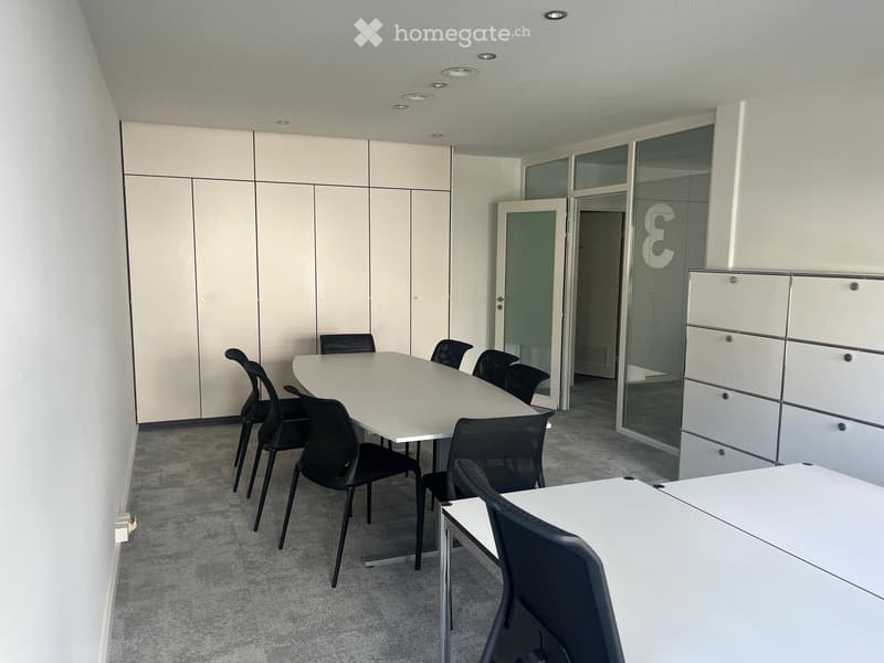 Modern office space with white walls, carpeted floor, white tables, and black chairs arranged around a central table.