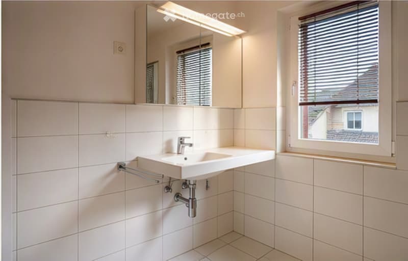 Modern bathroom with white tiled walls and floor, a single sink with a modern faucet, a large mirror above the sink, and a window with blinds.