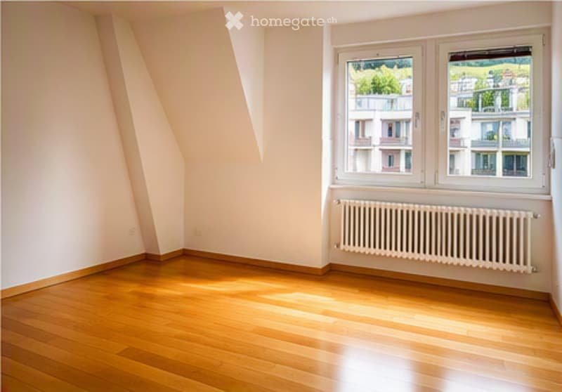Empty room with wooden flooring, white walls, and large windows showing a view of buildings outside. No furniture or equipment present.