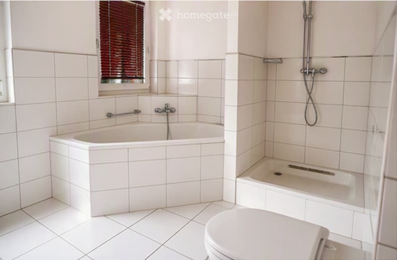 Bright bathroom with white tiled walls and floor, featuring a bathtub, toilet, and showerhead.