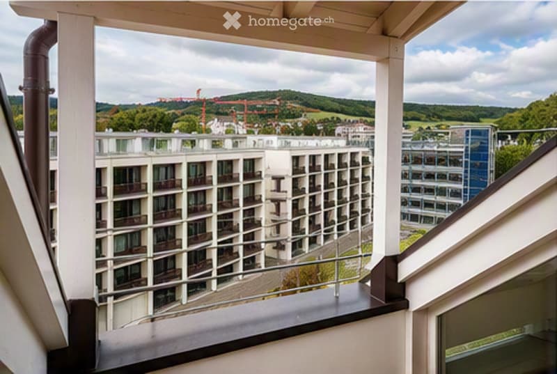 Balcony with a view of a modern apartment building and surrounding landscape, featuring white railings and a wooden floor.