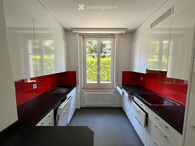 Modern kitchen with white cabinets, dark countertops, red backsplash, and a large window overlooking greenery.
