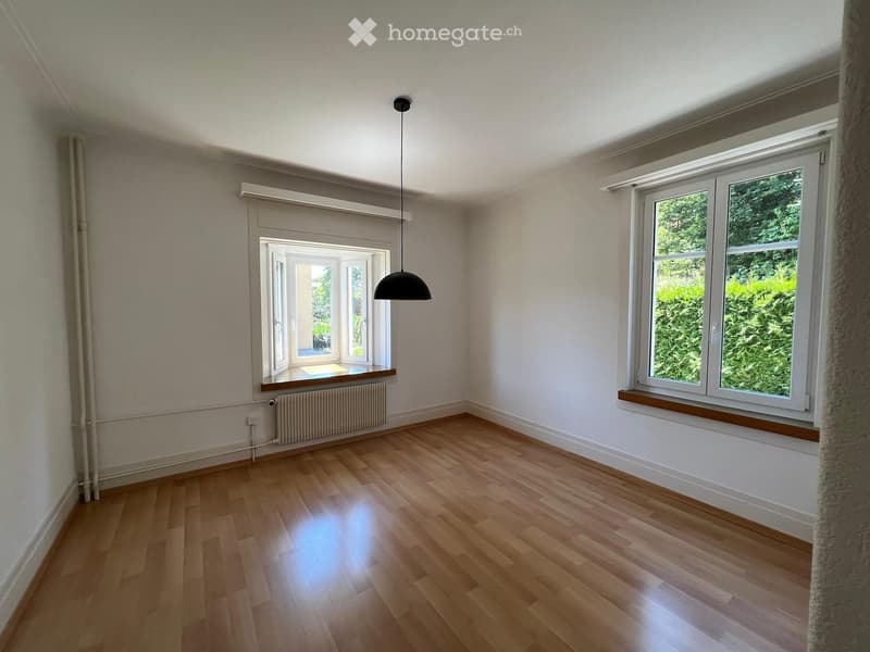 Empty room with wooden flooring, white walls, large windows, and a hanging light fixture.