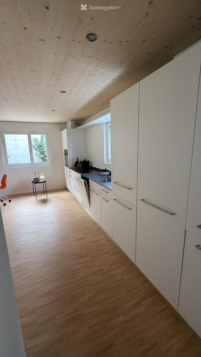 Modern kitchen with white cabinets, wooden flooring, and stainless steel appliances including a stove, oven, and dishwasher. A small table with a chair is near the window.
