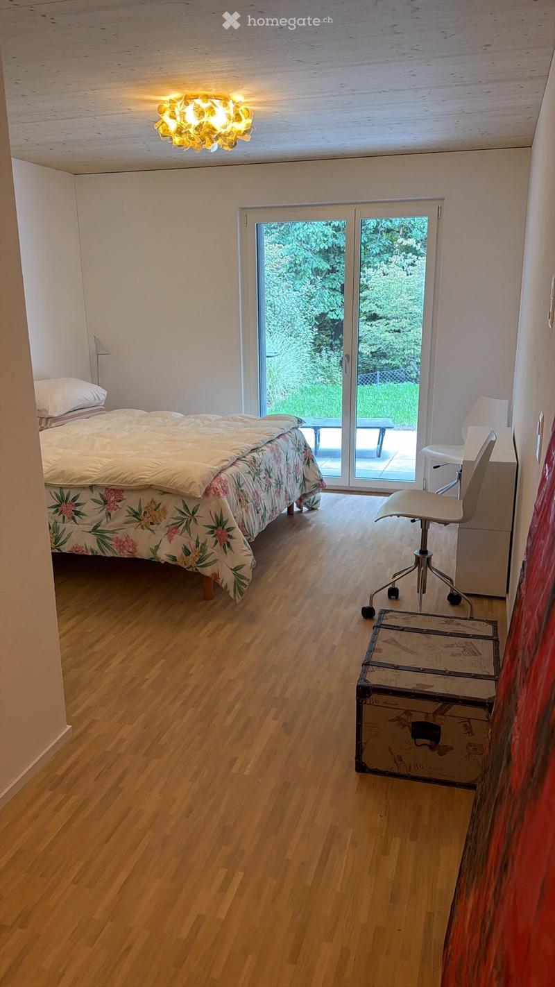 Cozy bedroom with wooden flooring, a bed with floral bedding, a desk with a chair, a lamp, and a suitcase near the desk. Large sliding glass doors lead to an outdoor area.