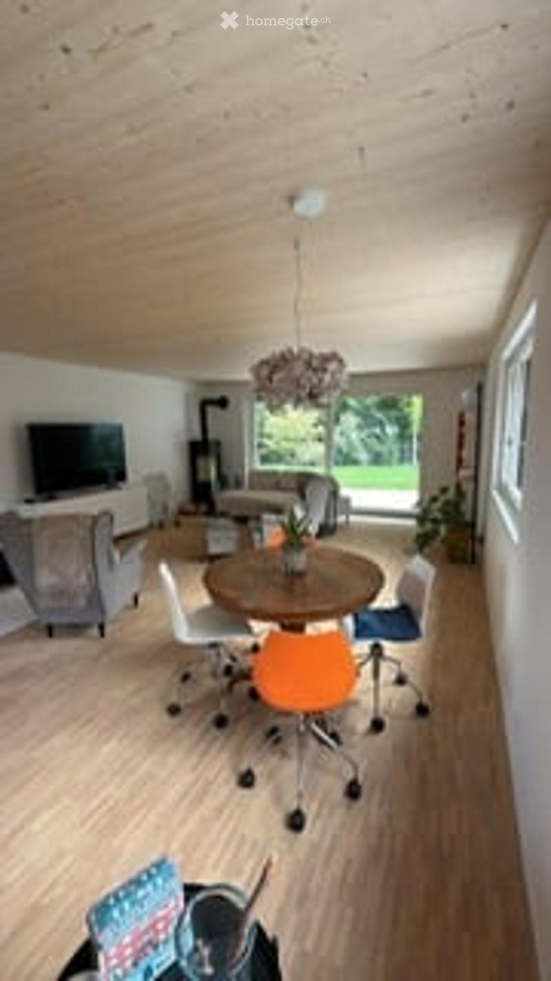 Bright, open-plan living and dining area with wooden flooring, large windows, a sofa, coffee table, dining table with chairs, and a television on a stand.