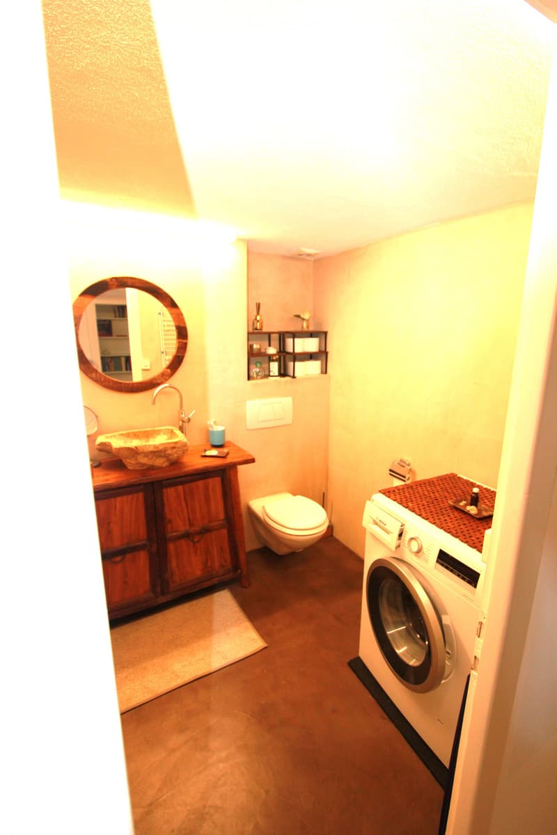 Modern bathroom with a wooden vanity, stone sink, round mirror, toilet, and a washing machine with a wooden countertop.