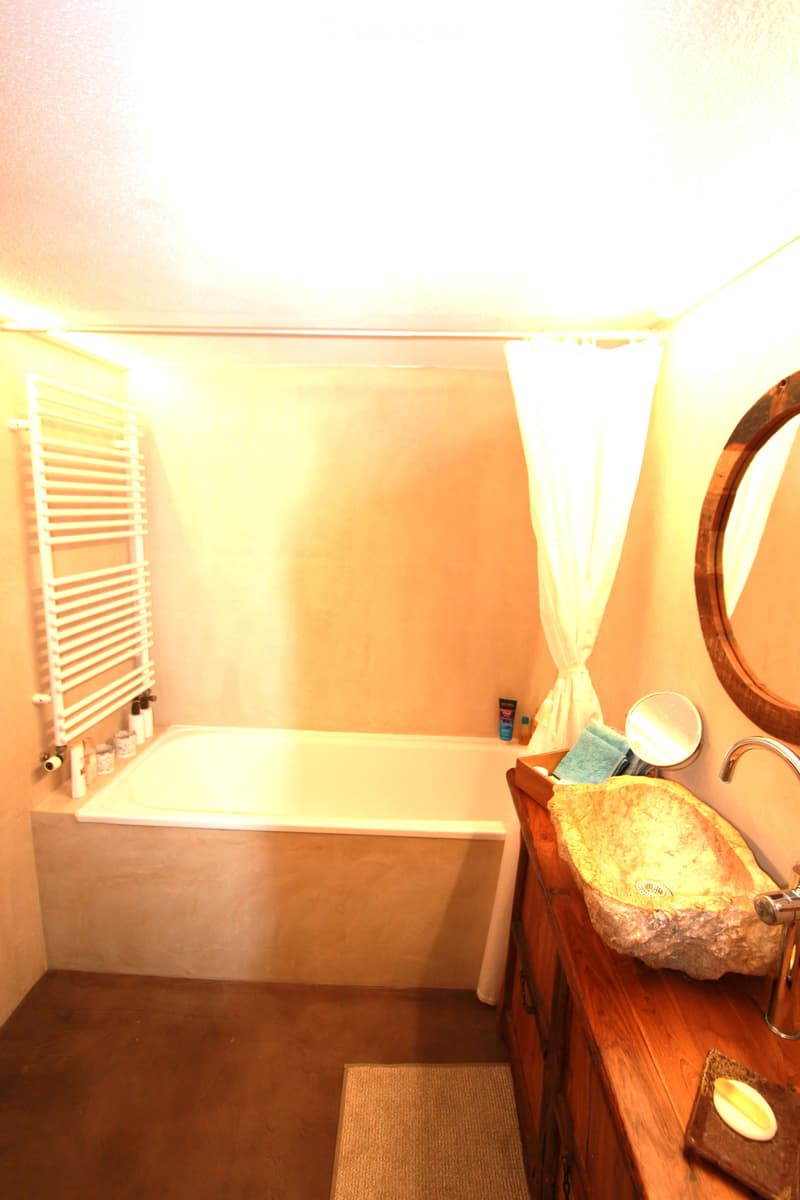 Bathroom with a bathtub, wooden sink, large mirror, and towel rack. The room features warm lighting and a wooden floor.